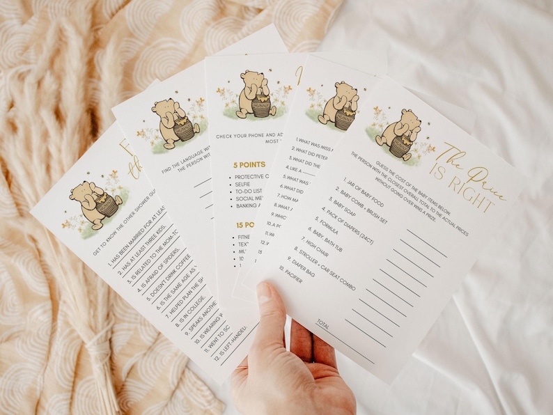 May include: Four baby shower game cards with Winnie the Pooh illustrations. The cards have gold and black text, with games like "The Price is Right" and "Get to Know the Other Shower Goer". The cards are on a cream-colored surface.