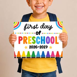 May include: A rectangular white sign featuring the text first day of PRESCHOOL 2026-2027 in black and multicolored lettering. The design includes colorful crayon illustrations along the bottom edge, rainbow arcs in the top corners, and yellow stars.