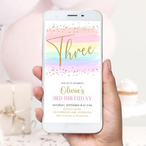 Puede incluir: Un teléfono inteligente muestra una invitación de cumpleaños. La invitación presenta la palabra "Three" en escritura dorada sobre un fondo de acuarela pastel. La invitación es para el tercer cumpleaños de Olivia, con detalles de fecha, hora y lugar.