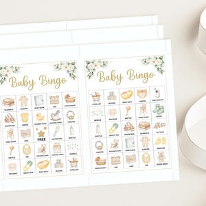 May include: Baby Bingo game cards with a floral design in the corners. The cards have a gold font that reads "Baby Bingo". The bingo cards have illustrations of baby-related items such as a stroller, blocks, and a baby bottle.