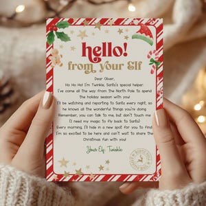 Puede incluir: Una tarjeta navideña, sostenida por manos, con un borde a rayas rojas y blancas. La tarjeta dice "hello! from your Elf" con ilustraciones festivas y un mensaje de "Twinkle" a "Oliver".