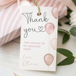 Può includere: Un'etichetta regalo con le parole "Thank you" in corsivo nero, con un simbolo a cuore. L'etichetta presenta palloncini rosa e il testo "For coming to my birthday party". Un regalo confezionato con strisce rosa e bianche è sullo sfondo.
