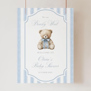 May include: A baby shower sign with a light blue and white striped background. The sign features a watercolor illustration of a teddy bear with a blue bow. Text reads "We can Bearly Wait" and "Welcome to Olivia's Baby Shower".