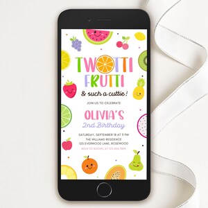 May include: A smartphone displays a colorful birthday invitation with the text "Twotti Frutti & such a cuttie!" and "Olivia's 2nd Birthday." The design features cartoon fruits like watermelon, strawberries, and kiwi. The phone is on a white surface.
