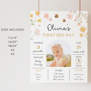 May include: A personalized 'First Bee-Day' sign featuring a baby's photo, floral and bee illustrations. The sign includes the baby's name, weight (21 pounds), height (28 inches), nicknames, and favorite things. Available in various sizes.