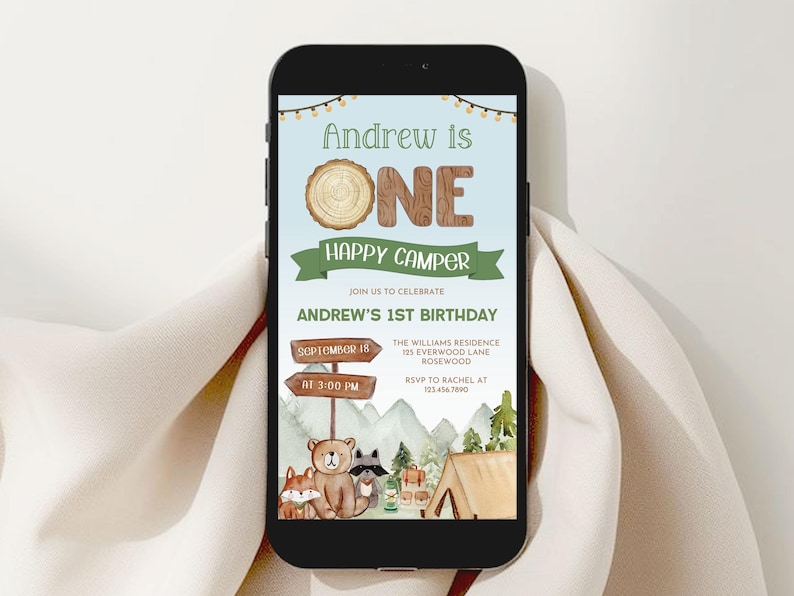 May include: A smartphone displays a digital invitation for Andrew's 1st birthday. The invitation features a camping theme with a bear, fox, and raccoon illustration. Text includes "Andrew is ONE Happy Camper" and event details.