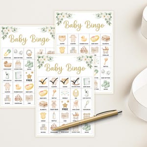 May include: Three Baby Bingo cards with watercolor illustrations of baby items like a rubber duck, crib, and bottle. The cards have a floral design and the words "Baby Bingo" in gold script. A gold pen and white ribbon are also in the image.