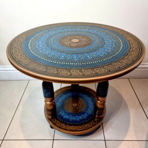 May include: Round wooden side table with a two-tiered design. The tabletop and lower shelf feature intricate blue and gold mandala patterns. The table has a brown frame and legs, with a dark blue and gold design on the legs.