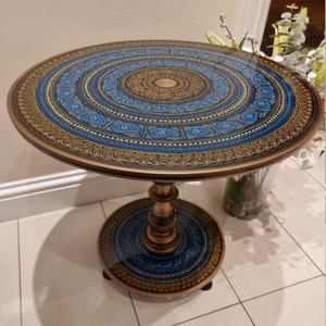 Op de afbeelding: Ronde bijzettafel met een gedetailleerd, sierlijk ontwerp. Het tafelblad heeft een blauw en goud mandala patroon. De tafel heeft een voetstuk en een bijpassende cirkelvormige basis. De tafel heeft een diameter van ongeveer 61 cm.