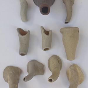 May include: A collection of ten ceramic pottery pieces, including a larger, brown, round-bottomed piece with a small opening at the top, and nine smaller, light brown, elongated pieces with a rounded opening at one end.