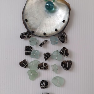 May include: A seashell wind chime with black and green glass pieces hanging from it. The seashell is white with black accents and a blue glass bead in the centre.