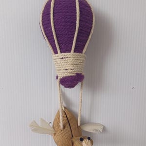 Könnte beinhalten: Ein lilafarbener Heißluftballon aus Garn mit einem beigefarbenen Korb aus Garn hängt von der Decke. Ein kleiner Holzvogel mit Flügeln aus Federn hängt am Ballon.