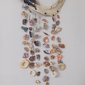 Op de afbeelding: Een windgong van drijfhout met een blauwe en zwarte veer die aan de bovenkant hangt. De gong is versierd met verschillende schelpen en stenen in tinten roze, oranje, bruin en wit. De schelpen zijn aan elkaar gebonden met kleurrijk touw.