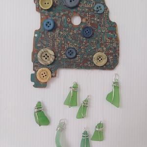 Puede incluir: Un carillón de vidrio verde con siete piezas de vidrio de mar envueltas en alambre plateado que cuelgan de una placa de circuito con varios botones de colores.