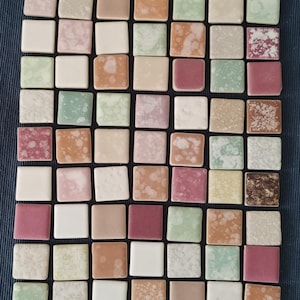 Puede incluir: Una colección de 64 pequeñas baldosas cuadradas de cerámica en varios tonos de rosa, beige, marrón y verde. Cada baldosa tiene un patrón moteado único.