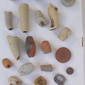 Könnte beinhalten: Eine Sammlung von 20 kleinen, handgefertigten Keramikperlen in verschiedenen Formen, Größen und Farben. Die Perlen sind aus Ton gefertigt und haben ein rustikales, erdiges Aussehen. Einige Perlen sind rund, andere oval und wieder andere zylindrisch. Die Perlen sind eine Mischung aus Braun, Beige, Grau und Rot.