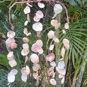 May include: A handcrafted wind chime featuring a collection of natural seashells in various shapes and sizes. The shells are primarily white, pink, and beige, suspended from a curved, rustic metal frame with pink twine accents. The background is lush green foliage.