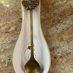 Op de afbeelding: Een witte keramische lepelsteun met een gouden rand en een bijendesign. Een gouden lepel met een bij-vormig handvat rust in de lepelsteun.