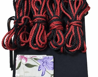 May include: A set of four coiled ropes featuring a twisted black and red pattern, resting on a solid black rectangular fabric base. A small, folded piece of light-colored fabric with a floral print in purple, pink, and green is placed on the base.