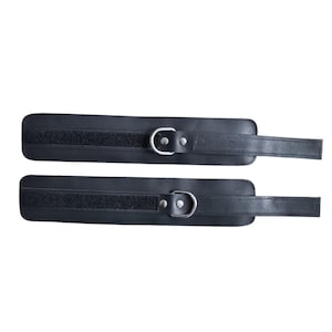 May include: Two black leather ankle restraints with silver-colored metal D-rings and hook-and-loop closures. Each restraint has a rectangular shape with a strap extending from one end.