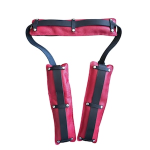 May include: Red leather restraint with black accents. The design features a padded headpiece connected to two vertical arm cuffs. Black straps and silver-toned rivets secure the pieces. The leather has a textured appearance.