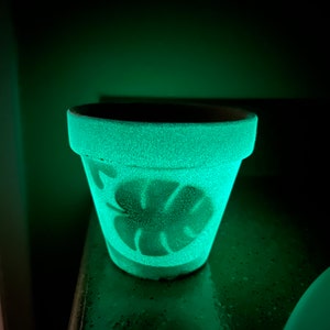 Puede incluir: Una maceta de terracota verde con un diseño de hoja blanca brilla en la oscuridad. La maceta está sentada sobre una superficie oscura.