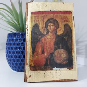 May include: A religious icon depicting an angel with a halo, holding a staff and a globe. The icon has a gold leaf border and is mounted on a wooden base. The angel wears a red and black robe. A blue, textured vase with a green plant is in the background.