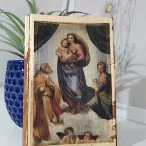 May include: A framed print of a Renaissance painting depicting the Madonna and Child with two angels and Saint Joseph. The painting is set against a gold background with a wooden frame.