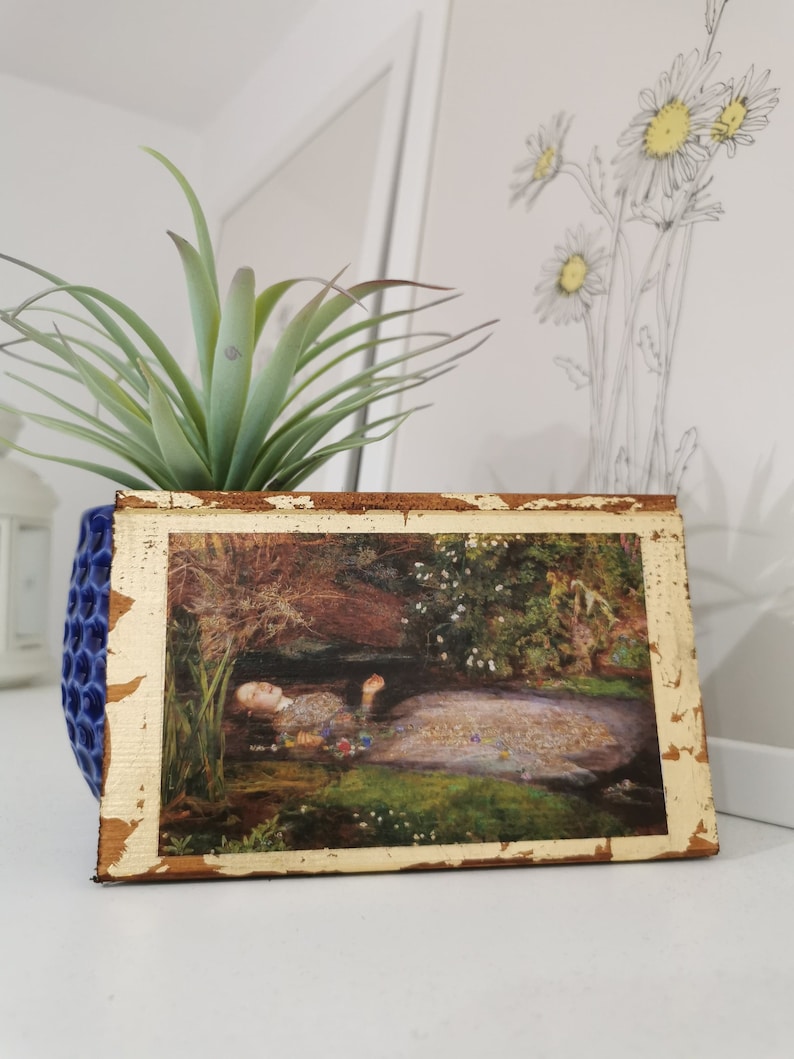 May include: A framed print of a painting depicting a woman lying in a stream of water surrounded by flowers and greenery. The frame is gold with a distressed finish.