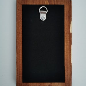 May include: A wooden frame with a black background and a silver hanging hook. The frame is made of dark wood with a light brown stain.
