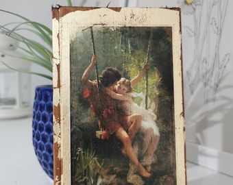 May include: A vintage wooden frame with a gold-painted border, featuring a painting of a couple on a swing in a forest setting. The painting is in a romantic style, with the couple embracing.