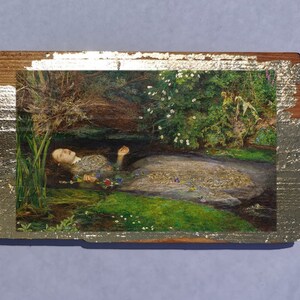 May include: A painting of a woman lying in a stream of water. The woman is wearing a white dress and has long, dark hair. The water is clear and the stream is surrounded by green plants and flowers.