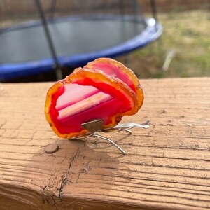 May include: A pink and orange agate butterfly with silver wire legs. The butterfly is sitting on a wooden surface.