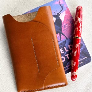 May include: A tan leather card holder with a curved pocket design and visible stitching, positioned next to a red and white marbled fountain pen and a notebook featuring a graphic illustration of a deer against a purple and pink mountain landscape.