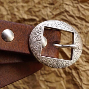 May include: A brown leather belt featuring a silver-toned oval buckle with intricate engraved scrollwork patterns. The belt is secured with a smooth silver-toned metal rivet, and the buckle frame is rectangular with rounded edges.