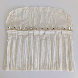 May include: A cream-colored, pleated fabric utensil roll with silver-colored silverware peeking out of the pockets. The roll is open, displaying the silverware. The fabric has a slightly wrinkled texture.