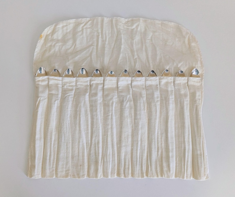 May include: A cream-colored, pleated fabric utensil roll with silver-colored silverware peeking out of the pockets. The roll is open, displaying the silverware. The fabric has a slightly wrinkled texture.