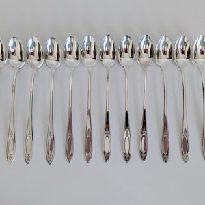 May include: A collection of eleven silver spoons arranged in a row. Each spoon has a long, slender handle and a rounded bowl. The spoons are made of shiny, reflective metal, and are likely for serving or eating.