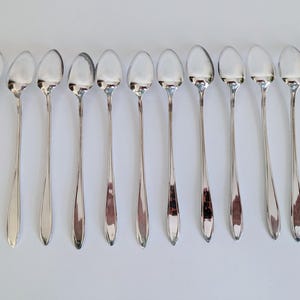May include: A collection of eleven silver-colored spoons arranged on a white surface. The spoons have long, slender handles and oval-shaped bowls. The spoons are evenly spaced and reflect the light, creating a shiny appearance. The spoons are likely for serving or eating.