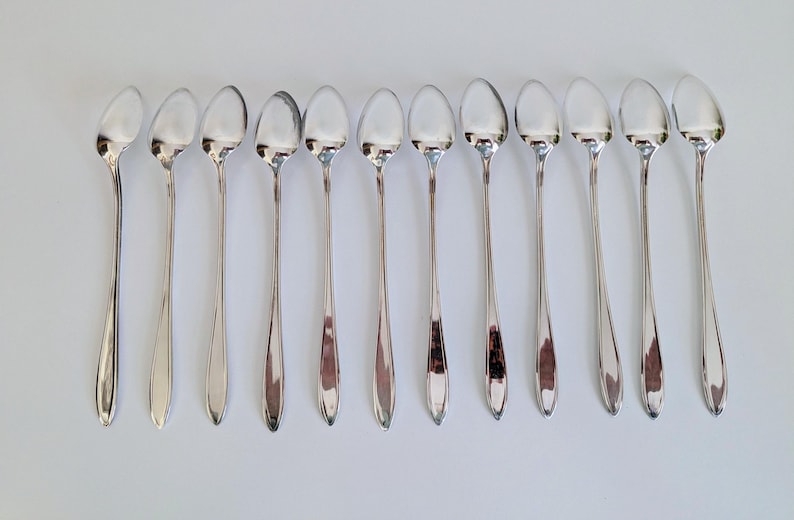 May include: A collection of eleven silver-colored spoons arranged on a white surface. The spoons have long, slender handles and oval-shaped bowls. The spoons are evenly spaced and reflect the light, creating a shiny appearance. The spoons are likely for serving or eating.