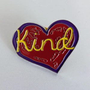 May include: A heart-shaped pin with a red center and a purple border. The word "Kind" is written in yellow cursive across the heart. The pin is a vibrant accessory.