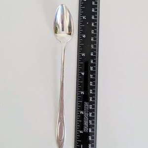 May include: A silver spoon with an oval bowl and a long, slender handle. The handle features a decorative design near the base. The spoon is approximately 7 inches long, next to a black ruler with inch measurements.