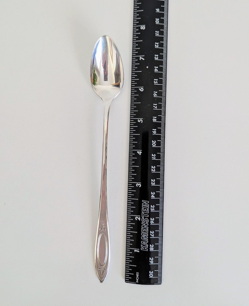 May include: A silver spoon with an oval bowl and a long, slender handle. The handle features a decorative design near the base. The spoon is approximately 7 inches long, next to a black ruler with inch measurements.