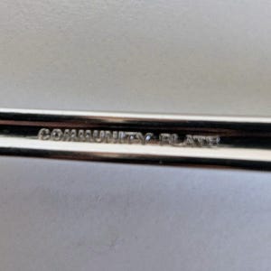 May include: Close-up of a silver utensil with the text "COMMUNITY PLATE" engraved on the handle. The utensil has a shiny, reflective surface, suggesting it is made of metal. The background is a light, neutral color.