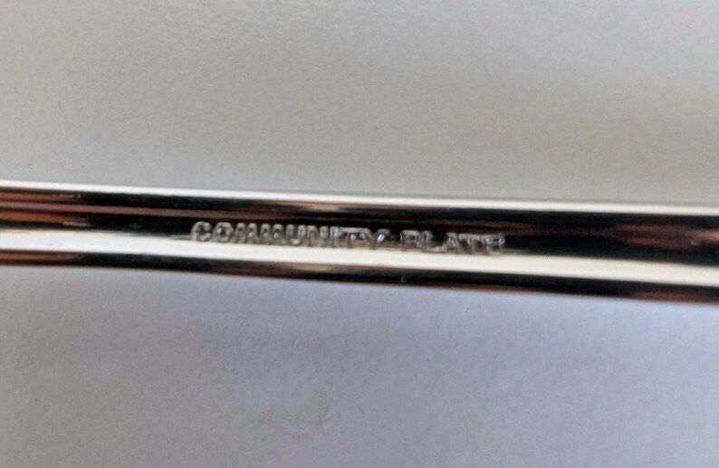 May include: Close-up of a silver utensil with the text "COMMUNITY PLATE" engraved on the handle. The utensil has a shiny, reflective surface, suggesting it is made of metal. The background is a light, neutral color.