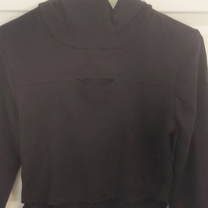 May include: A black cropped hoodie with a high, folded collar and long sleeves. The hoodie has a horizontal slit across the chest and a raw, unfinished hemline. The garment is hanging on a white hanger against a white background.