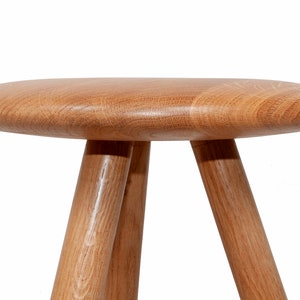 May include: A wooden stool with a round, flat top and three legs. The stool is made of light brown wood and has a natural finish.