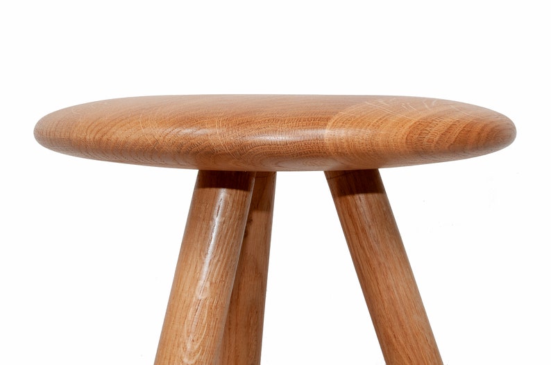 May include: A wooden stool with a round, flat top and three legs. The stool is made of light brown wood and has a natural finish.