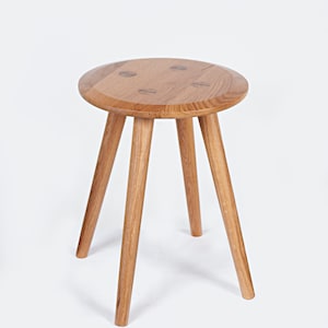 Puede incluir: Un taburete de madera con una parte superior redonda y tres patas. El taburete está hecho de madera marrón claro y tiene un acabado natural.