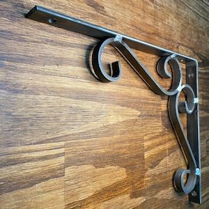 Puede incluir: Un soporte de estante de metal con un diseño decorativo de desplazamiento. El soporte está hecho de metal oscuro y tiene un acabado rústico.